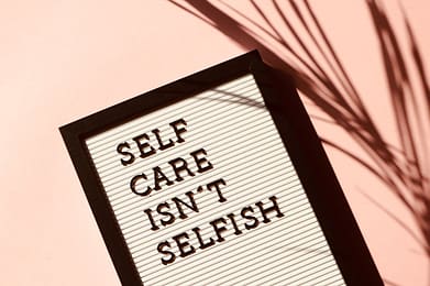 hero-img-01 Letter board displaying 'Self Care Isn't Selfish' with artistic shadow on pink background.